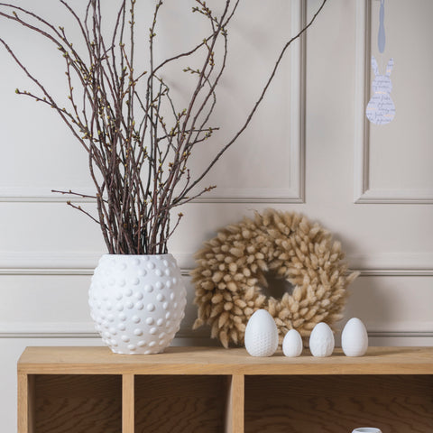 Decorative setup with a textured white vase, branches, and porcelain white easter eggs on a wooden surface.