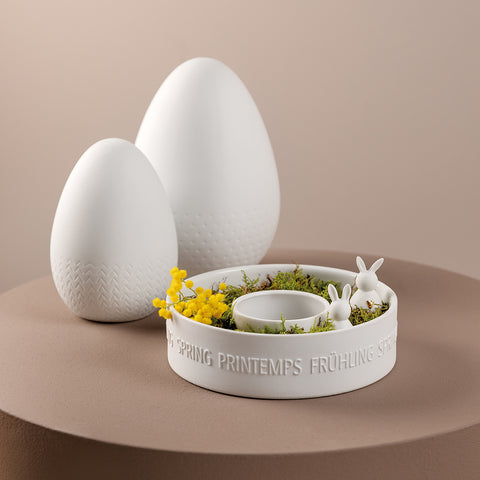Two white porcelain bunnies in a container with decorative eggs in the background.