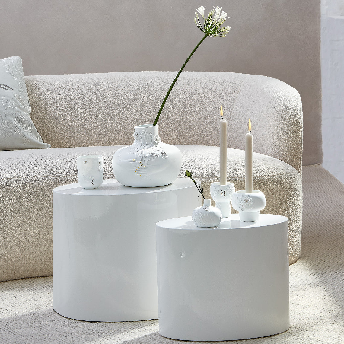 White porcelain vases and candle holders on a round white table with a beige sofa in the background.