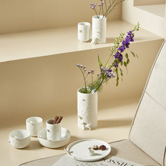 Porcelain Stories white porcelain tableware set with a vase of purple flowers on a beige surface.