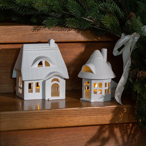 Two white ceramic house-shaped lanterns on a wooden surface with Christmas decorations.